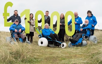 Beach wheelchair volunteers holding £1000 sign in the sand dunes